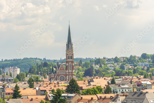 View of Herz Jesu Kirche soaring above a tapestry of red-tiled roofs, nestled amidst lush greenery under a soft sky, Herz jesu kirche church, Graz, Austria.