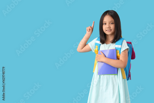 Cute little happy Asian girl with backpack and notebooks pointing at somethin...