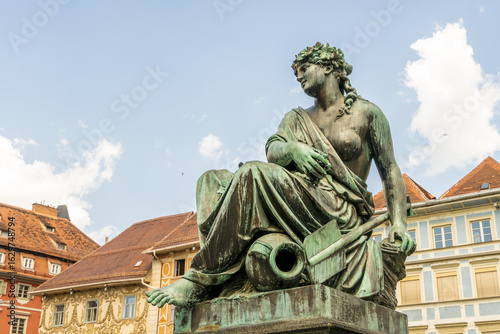 View of a bronze statue of a woman with a jug and an oar sits regally against a backdrop of warm terracotta roofs and a blue sky, Graz, Graz, Austria.