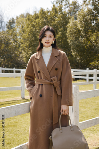 Woman in a brown coat standing near a fence.