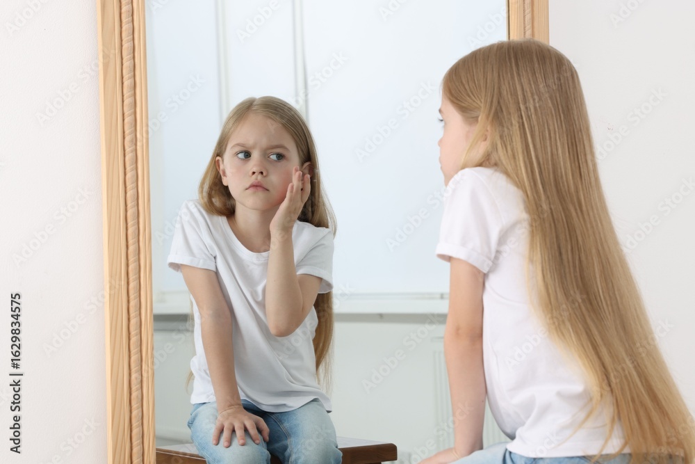 Fototapeta premium Upset little girl looking at herself in mirror indoors