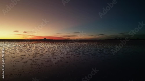 Drone footage moving forward over Salar de Uyuni salt flats at dusk. Clear sky transitions from deep blue to warm golden hues near horizon. Mountain silhouettes visible in distance.