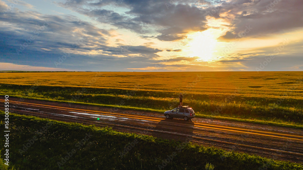 Fototapeta premium a man on the roof of a car in a wheat field at sunset