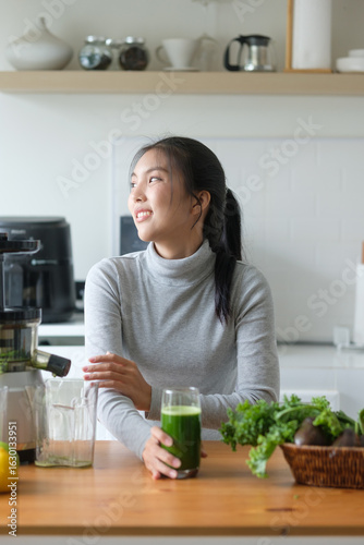 Young Asian woman holding a glass of green juice, smiling and relaxing in a modern white kitchen.