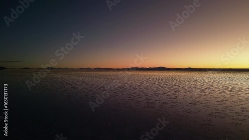Drone footage moving forward over Salar de Uyuni salt flats at dusk. Clear sky transitions from deep blue to warm golden hues near horizon. Mountain silhouettes visible in distance.