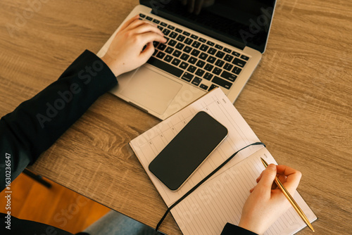 Person working at wooden desk with laptop, writing in a notebook with golden pen, smartphone placed on notebook, modern office environment