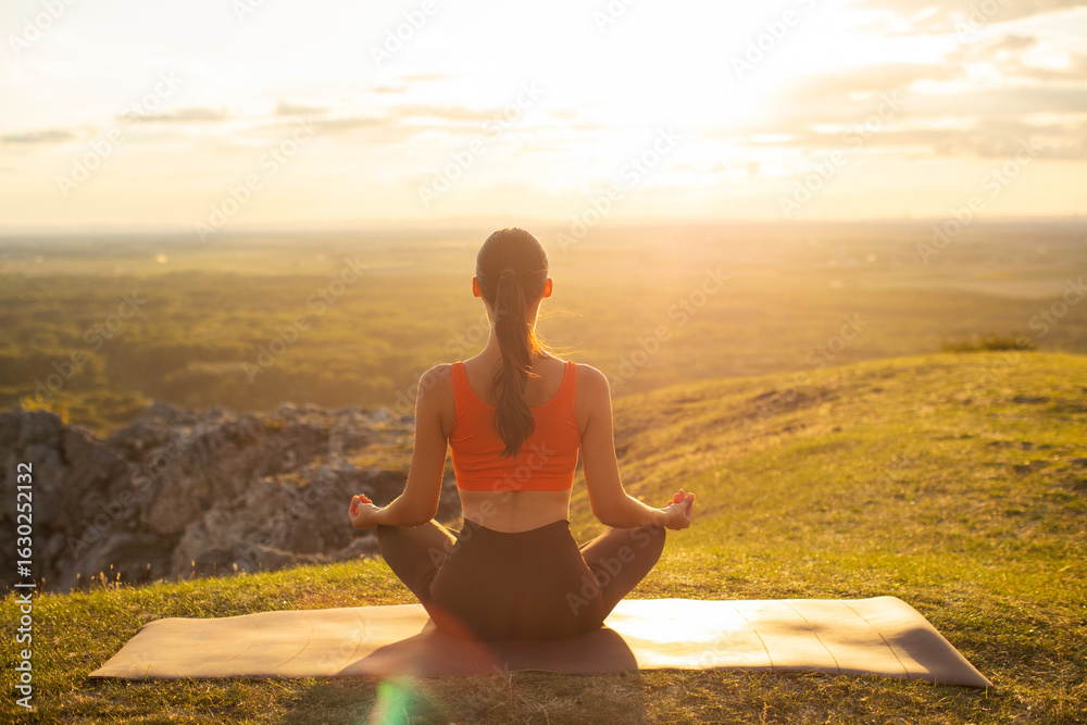 Fototapeta premium Back view of woman meditating in yoga asana in the mountains at the sunset outdoors. Spiritual practice concept