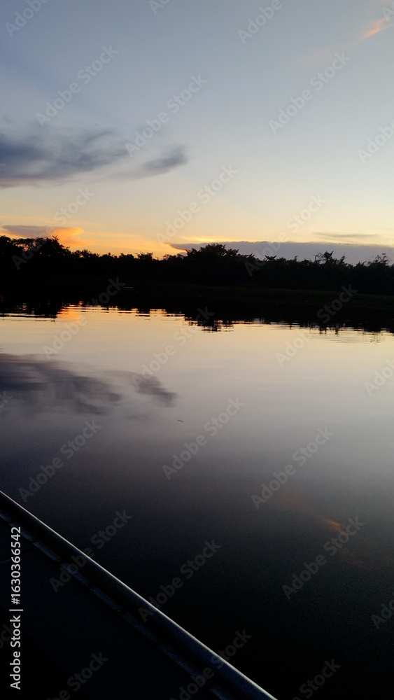 Obraz premium Cuiabá River in Pantanal wetlands at dusk with orange sunset reflecting on the calm water surface. A dark silhouette of the forest on the horizon under a twilight sky. Peaceful natural landscape view.