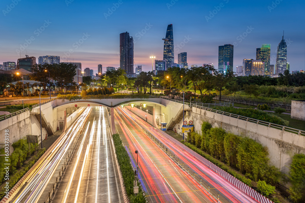 Obraz premium Urban night scene of Ho Chi Minh City featuring modern skyscrapers in the background and a green park surrounding a busy underpass in the foreground