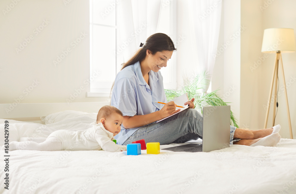 Fototapeta premium Cheerful smiling happy young mother in casual working on a laptop computer while her cute little child playing with toys lying on bed in bedroom at home. Motherhood and education concept.