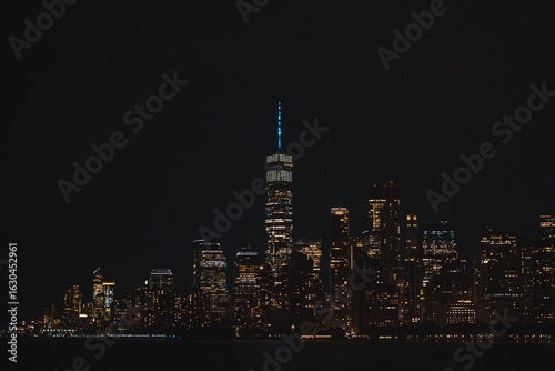 The illuminated Lower Manhattan skyline and One World Trade Center glittering at night, seen from the water.