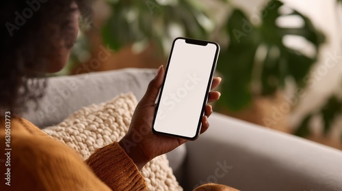 young black african woman holding smartphone in hand with white black empty mockup screen template using cell phone relaxing on sofa at home over shoulder view mobile apps advertising concept no logo