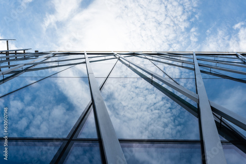 Tall modern building with glass facade and metal beams reflecting clouds and blue sky, viewed from below. Suitable for illustrating architecture, urban development, or corporate design concepts.