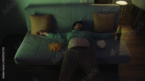 A middle-aged adult man with a bare belly and in home clothes lies and sleeps on the sofa at night with chips, popcorn and a bottle in front of the TV in room. A lazy person tired from work