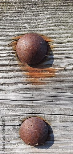 Close-up of two old rusted metal rivets embedded in aged wooden planks, with visible rust stains and cracked wood texture under sunlight