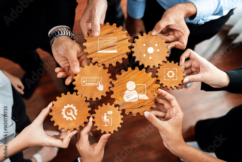This image showcases a collaborative effort with hands holding wooden gears that symbolize teamwork and innovative ideas in a modern office environment. Amity