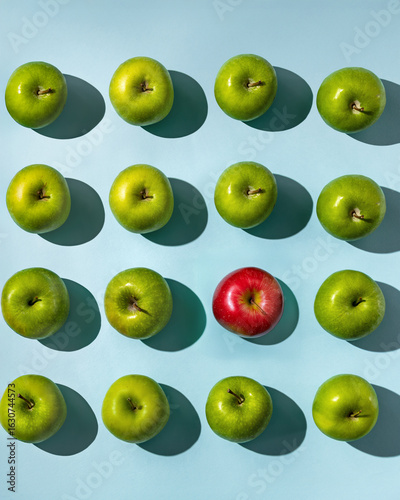 Фотография One red apple standing out in a group of green apples on a blue background