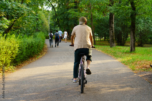 Wallpaper Mural Man rides bicycle along park path on sunny day, surrounded by greenery and other park-goers Torontodigital.ca