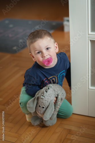 Fotografie A young boy with a pacifier poses indoors on a wood floor
