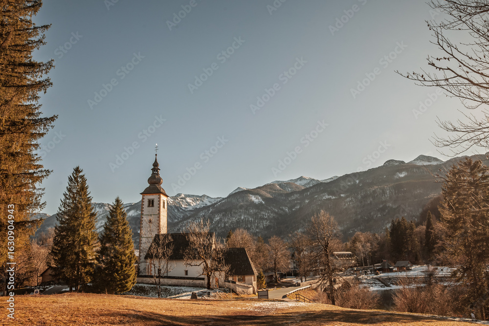 Fototapeta premium Church of Saint John the Baptist - Cerkev svetega Janeza Krstnika - at Ribcev Laz by Lake Bohinj, Slovenia, photographed at dusk with snowy peaks and village houses in an alpine scene.