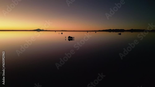 Drone footage circling 4x4 vehicle and two people on chairs at salt flats during sunset. Perfect mirror reflection on water surface. Mountains silhouetted on horizon at dusk.