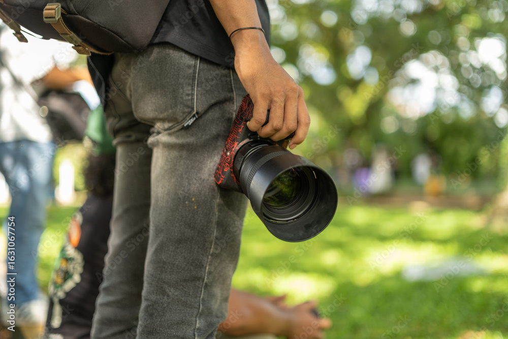 Fototapeta premium Close-up of a man holding a DSLR camera with a telephoto lens in a park, captured in natural daylight with a blurred green background, conveying photography lifestyle and outdoor action.