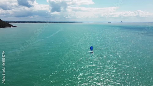 Drone footage follows a sailboat sailing across the coastal waters of Normandy, with Mont Saint-Michel appearing slowly on the horizon.
