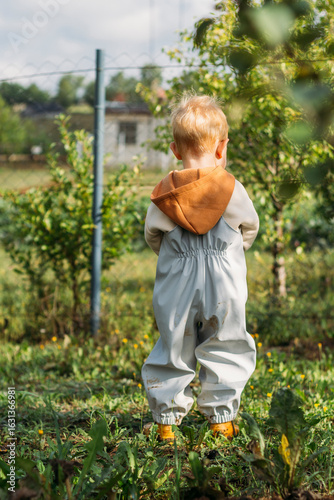 A small red-haired boy in a gray-blue jumpsuit looks into the distance over the fence