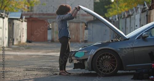 Woman looks under the hood of her car, he is upset and annoyed by the breakdown trying to repair herself