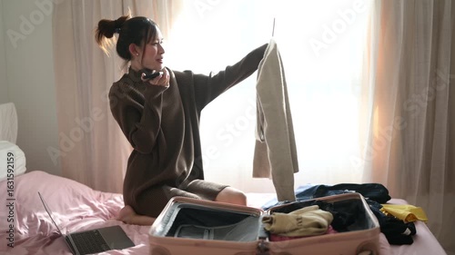 A cheerful woman sits on a bed, packing a suitcase while holding up a sweater, surrounded by clothing and a laptop.