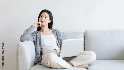 Free Download A thoughtful and focused young woman working on her laptop while sitting comfortably on a gray sofa, portraying a modern and flexible remote work lifestyle. - GFXLoad