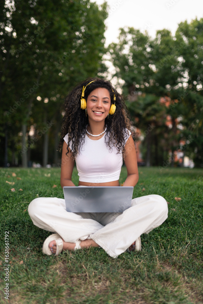 Obraz premium Smiling young woman working with a laptop and headphones in a park surrounded by many trees