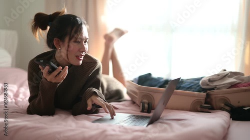 A woman is lying on a bed, smiling while talking on her phone and using a laptop, with luggage and clothes visible nearby.