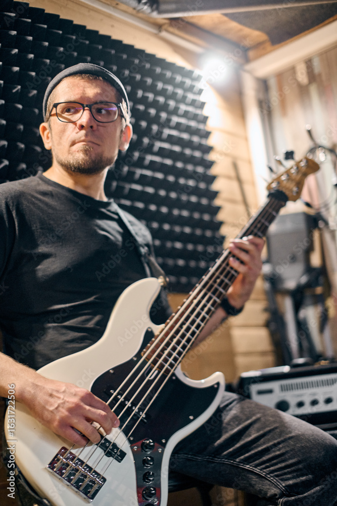 Fototapeta premium Young male musician with glasses and beanie plays electric bass guitar in wood-paneled studio. Soft lighting and soundproof panels create focused atmosphere