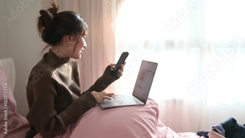 A young woman sits on a bed, holding a smartphone, with soft morning light filtering through a window, creating a serene atmosphere.