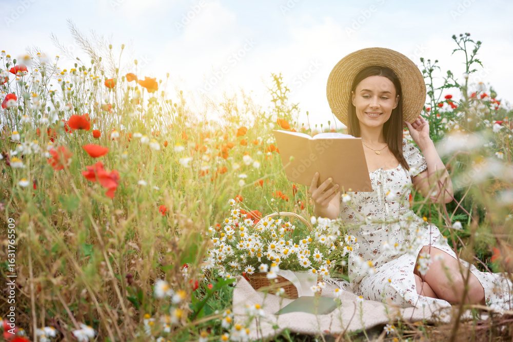 Fototapeta premium Smiling woman in hat reading book in morning wildflower meadow