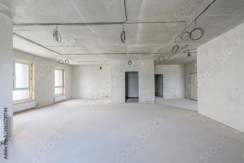 spacious, empty room under construction. Cement walls and floor, exposed wiring on the ceiling, and window openings await finishing