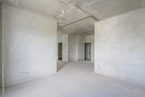 An unfinished, empty room with concrete walls and ceiling. Visible wiring and doorways suggest ongoing construction or renovation