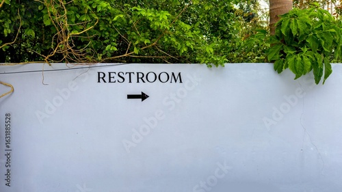A close-up shot captures a minimalist restroom sign with a black arrow pointing to the right, painted on a clean, light-colored wall. The upper edge of the wall is bordered by lush, green tropical