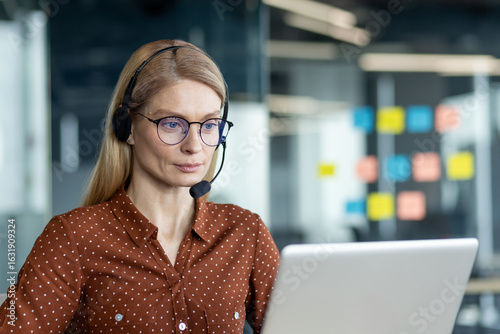 Photography A female customer service representative wears a headset while working on her laptop in a modern office setting