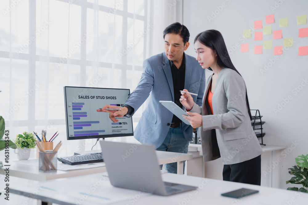 Naklejka premium Two asian businesspeople analyzing financial charts displayed on a computer screen, discussing strategies and collaborating on a project in a modern office environment