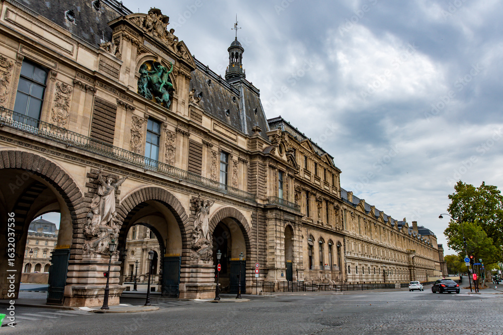 Fototapeta premium Stunning perspective of the Louvre Museum's historic architecture and the street life below under a cloudy sky.
