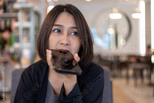 Canvas-taulu Asian woman sitting on couch at cafe hand holding mobile phone near mouth using