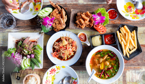Top view of a colorful Thai meal with traditional dishes, fresh vegetables, fried chicken, papaya salad, spicy soup, and dipping sauces on a rustic wooden table.
