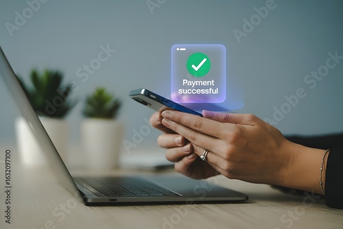 Close up of hands holding a smartphone displaying a successful payment confirmation with a green checkmark overlayed on a laptop screen in a modern office setting