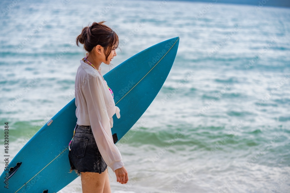 Fototapeta premium Young woman walking along the beach with a blue surfboard on a sunny day, preparing for a surfing session near the ocean