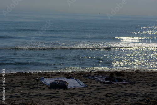 Photography Two towels and a pair of shoes are seen left on a sandy beach next to the calm sea on a sunny day