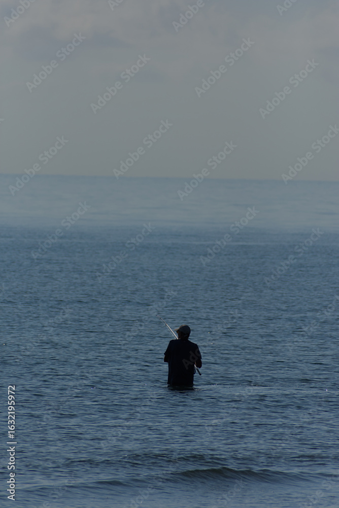 Fototapeta premium A lone man is seen standing in the sea, fishing with a long rod on a calm, misty day.