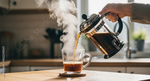 A person's hand carefully pours steaming hot coffee from a classic French press into a clear glass mug on a wooden kitchen counter, creating a cozy morning scene.