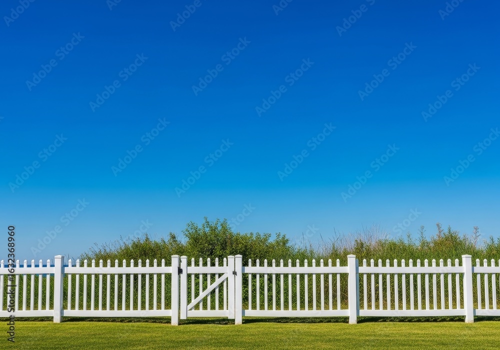Fototapeta premium Peaceful White Fence Under Blue Sky - Serene landscape, white picket fence, vibrant green grass, clear blue sky, symbolizes peace, tranquility, and simple beauty